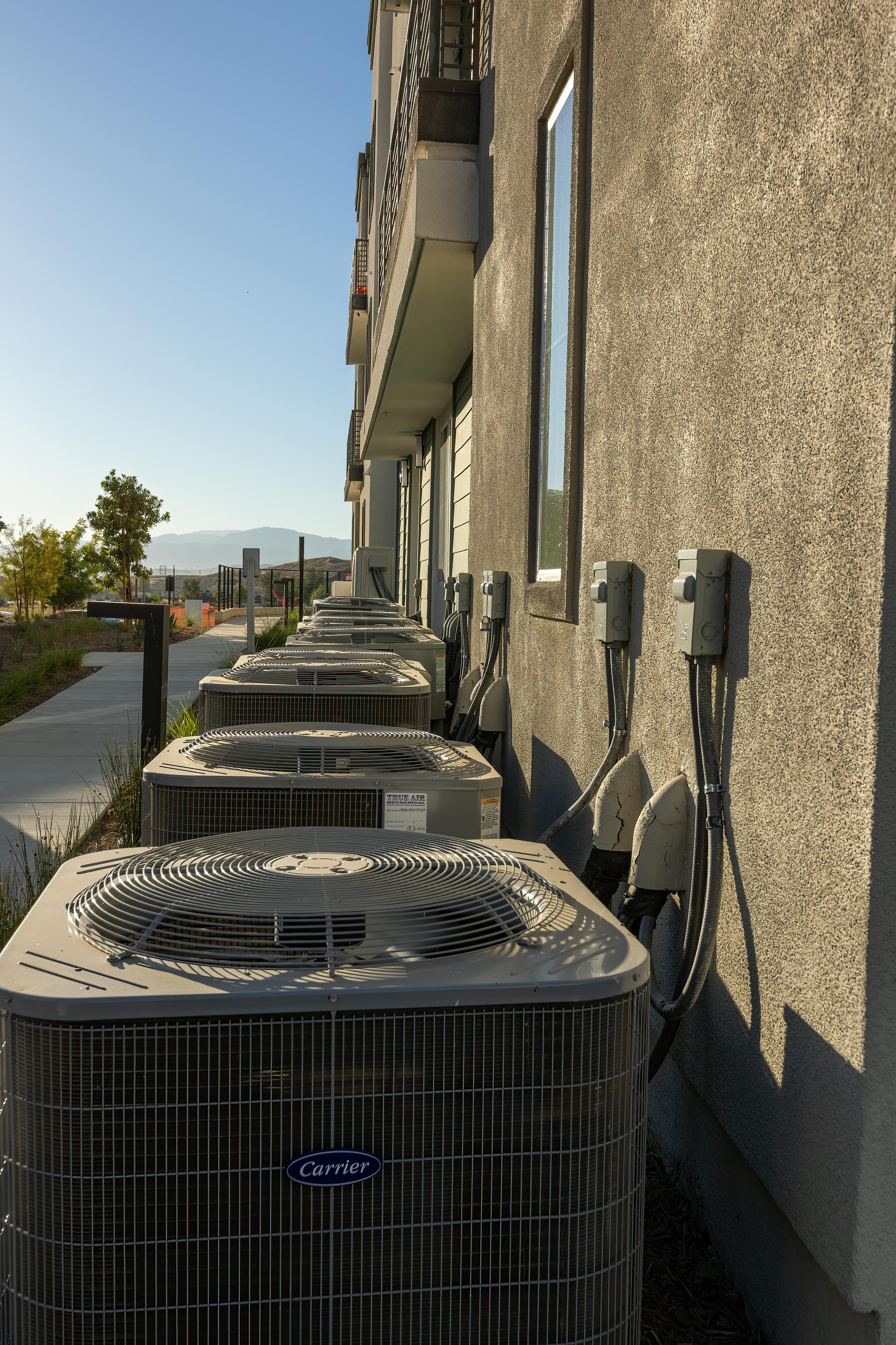 Line of air conditioning units on a city apartment exterior with a sidewalk view.